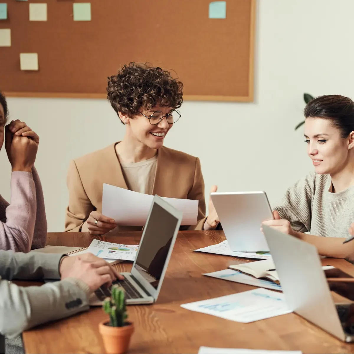 A group of professionals engaged in a discussion around a table, with laptops, documents, and a small plant in a collaborative setting.